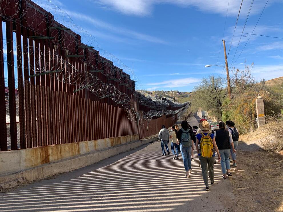 A group of people walk along a very tall metal fence with barbed wire across the top.