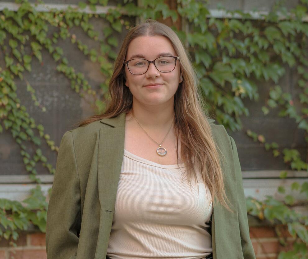 Ashley stands in front of a wall of ivy, smiling and wearing a green blazer and cream-colored shirt. 