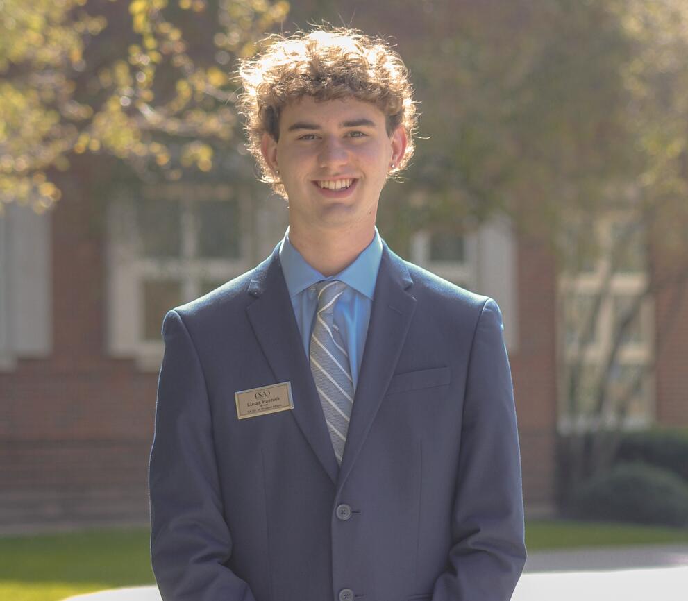 Lucas stands in front of South hall, wearing a blue blazer and striped tie. 