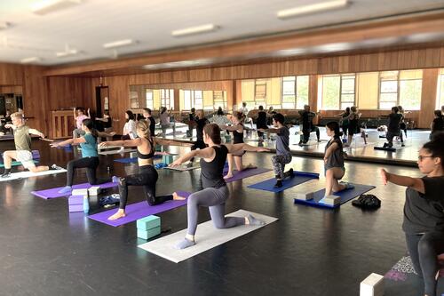 Approximately 12 students doing a yoga pose in a kneeling position on yoga mats in the Schrader Dance Studio, where this are mirrors in the background