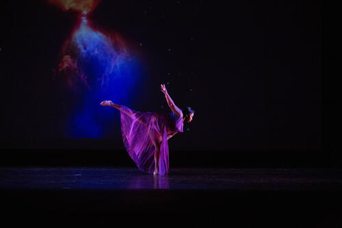 A dancer leans forward on stage with blue fabric fanning behind her.