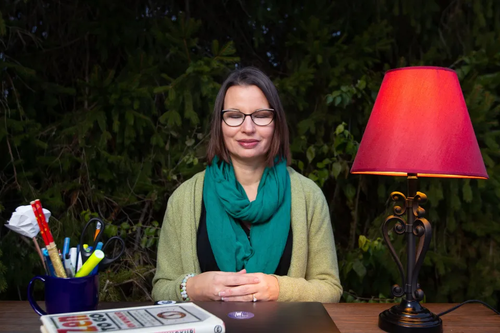 Beth Cholette sitting in front of a desk that has a lamp with a red shade, books, and pens