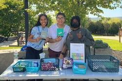 Table with baskets of information; three students are student behind the table, with the one on the far right wearing a mask; it is outdoors with trees in the background