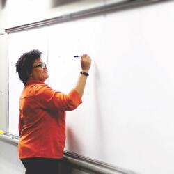 A smiling Black woman with natural hair and an orange shirt writes on a whiteboard